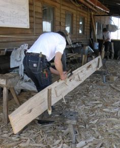 Mike then uses a broad axe - with a long, straight cutting edge - to slice through the partially-cut wood fibres. He moves the axe downwards, cutting from the top of the plank. This removes large shavings which are scattered on the ground. The axe handle curves to the right, away from the plank, so that Mike's hands pass freely with each stroke and don't hit the wood.